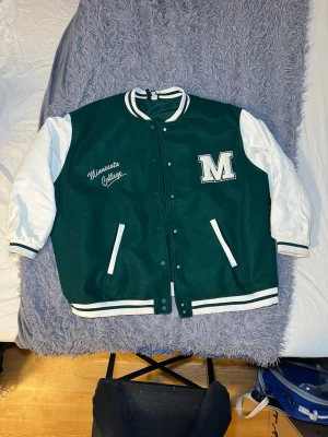 H&M+ BASEBALL JACKET Mörkgrön/Minnesota - Säljer en grön och vit bomberjacka från H&M med broderad text 'Minnesota College' och stor M-patch på bröstet. Jackan har vita ärmar, grönt kroppsparti, ribbade muddar och knappar framtill. Materialet är polyester och viskos, perfekt för dig som gillar college-stil. Nypris 499kr