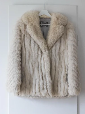 Saga Fox pälsjacka i ljus beige - Fluffig och lyxig pälsjacka från Saga Fox.  Jackan har bred krage, krokknäppning, samt ett klassiskt snitt och mjukt foder på insidan. Perfekt för dig som vill sticka ut med en statement piece under kalla dagar.