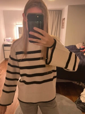 Randig stickad tröja i beige och svart - Säljer en snygg stickad tröja med breda svarta ränder på beige botten. Tröjan har rund hals, långa ärmar och en loose passform som ger en chill vibe. Perfekt att slänga på sig när det är lite kyligt ute.