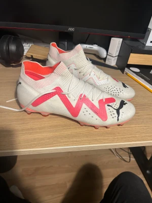 Fotbollsskor Puma FUTURE PRO FG/AG - Säljer ett par snygga Puma Future PRO fotbollsskor i vitt med rosa/röda detaljer. De är i storlek 44 och i princip i nyskick, använda 2-3 gånger och det ända som syns är lite smuts från konstgräs på undersidan av skon. Pris går att diskutera
