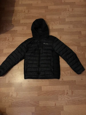 Svart pufferjacka från Champion S - Svart pufferjacka från Champion i storlek S. Jackan har huva, dragkedja framtill och en bröstficka med dragkedja. Quiltad design och stilren look, perfekt för kalla dagar. Materialet är syntet och jackan har långärmade ärmar.