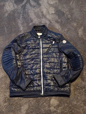 Moncler puffer cardigan - Snygg mörkblå pufferjacka från Moncler med quiltad design och vita dragkedjor. Jackan har ståkrage, långärmade ärmar och Moncler-logga på vänster arm. Materialet är glansigt och tillverkat i polyester så ett tunnt material, med puffer. Den har inga skador på sig. Perfekt för dig som vill ha en stilren och grisch stil. Vid köp gratis marmor armband!.