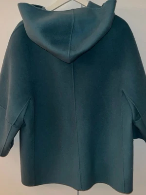 Turkos Max mara   kappa med huva  - Säljer en stilren turkos kappa från Max mara   med oversized passform och huva. Jackan har knappar framtill och är tillverkad i ett mjukt ullmaterial. Perfekt för dig som gillar minimalistisk design och vill ha en jacka som sticker ut med sin coola form.