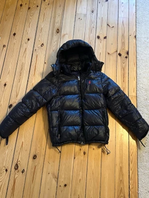 Polo Ralph Lauren puffer jacket - Svart pufferjacka från Polo Ralph Lauren med huva och tvåvägsdragkedja. Jackan har ett glansigt yttermaterial i polyester och är dekorerad med det klassiska röda Polo-logot på bröstet. Flera fickor med dragkedja och quiltad design för extra värme. Den har en liten defekt på ena armen men de är inget som syns så mycket. Hör av er vid frågor eller funderingar 