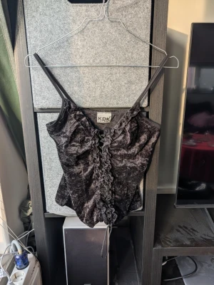 Svart sammetstopp med snörning KDK London - Säljer en svart croppad topp från KDK London i mjuk sammet. Toppen har smala axelband och snygg snörning framtill med rynkade detaljer längs mitten. Perfekt för dig som gillar en edgy och trendig look.