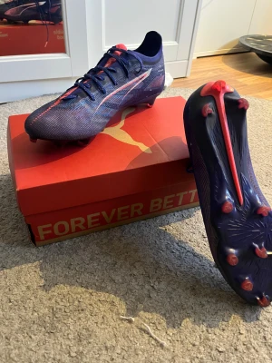 Blå fotbollsskor från Puma - Säljer ett par snygga fotbollsskor från Puma i mörkblå färg med röda och vita detaljer. Skorna har snörning och en spetsig tå, samt platt sula med röda dobbar för gräsplan. Ovandelen är tillverkad i syntetmaterial som ger lätt känsla och bra passform. Perfekt för dig som vill ha speed på planen.