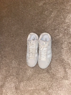 Adidas Campus vita sneakers - Säljer ett par fräscha Adidas Campus sneakers i vitt med klassiska tre ränder på sidan och guldfärgad logga bak. Skorna har snörning, platt sula och är gjorda i mocka och skinn. Perfekta för dig som gillar stilrena och tidlösa sneakers.