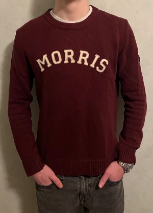 Vinröd stickad tröja från Morris - Snygg vinröd stickad tröja från Morris med vit broderad logga på bröstet. Rund halsringning och ribbade muddar vid ärmslut och nederkant. Perfekt för dig som gillar klassisk stil med en modern touch.