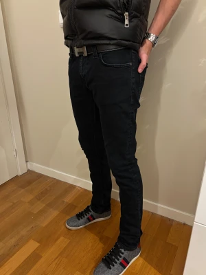 Nudie jeans - Säljer ett par svarta slimfit jeans från Nudie Jeans med klassisk femficksdesign och svart läderpatch bak i midjan. Jeansen har en smal passform och är tillverkade i stretchig bomull för extra komfort. Perfekt för dig som gillar en stilren och modern look.