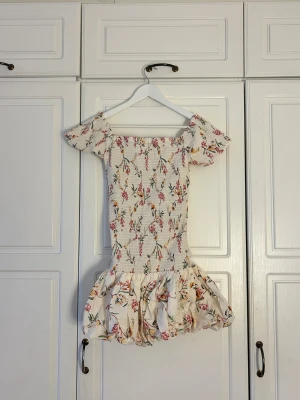 Blommig kortklänning med volang💖💖 - Supersöt kortklänning med offshoulder och puffiga ärmar. Klänningen är vit med blommigt mönster i rosa, gul och grön. Smockad överdel och volangig kjol ger en romantisk vibe. Perfekt för sommarens alla tillfällen!