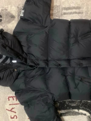 Svart burberry jacka  - Svart pufferjacka från Burberry London England med huva och dragkedja samt knappar framtill. Jackan har en quiltad design och en ficka med dragkedja på bröstet. Insidan har ett snyggt rutigt mönster i grått och svart. Perfekt för kalla dagar.