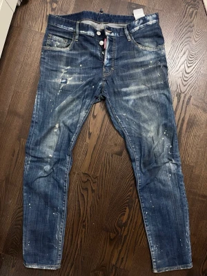 Dsquared2 blå jeans (äkta) - Säljer ett par blå jeans från Dsquared2 med coola slitningar och färgstänk. Jeansen har klassisk femficksdesign, knappgylf och tydliga detaljer som förstärkta sömmar och patch baktill. Materialet är robust denim och passformen är rak med något tapered ben. 