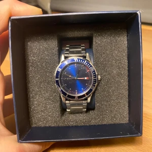 Blå klocka från Tommy Hilfiger - Säljer en snygg pojk klocka från Tommy Hilfiger med blå urtavla och silverfärgat armband i metall. Klockan kommer i originalbilden och 4 extra länkar medföljer. Just nu är den inre omkretsen cirka 14 cm utan länkar