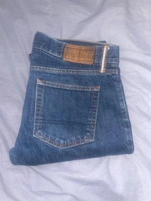 Blå  jeans storlek 31x30 från Tommy hilfiger  - Snygga blå jeans med klassisk rak passform och kontrastsömmar. Modellen har fem fickor och ett brunt läderpatch vid linningen. Perfekt för dig som gillar en enkel och tidlös stil. Materialet är slitstarkt jeans och färgen är mörkblå. Hör av er vid frågor, kan gå ner i pris vid snabb affär