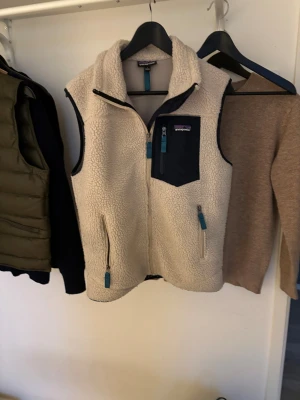 Beige fleeceväst från Patagonia - Säljer en beige fleeceväst från Patagonia i storlek S. Västen har marinblå detaljer, dragkedjor och en praktisk bröstficka. Materialet är mjuk fleece och västen har två sidofickor med dragkedja. Perfekt för lager-på-lager under kyliga dagar.