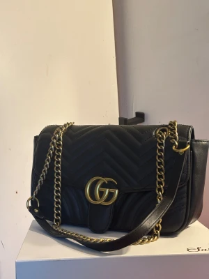 Svart  axel väska GG logga - Säljer en svart quiltad axelväska  med GG-logga i guld framtill och kedjerem i antikguld. Väskan har ett snyggt hjärtdetalj på baksidan och är tillverkad i mjukt skinn med chevronmönster. Perfekt storlek för det viktigaste och riktigt trendig look.
