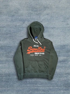 Superdry Hoodie - Grön hoodie ifrån Superdry. Tröjan är i bra skick men har några bristningar i den oranga texten. Annars är tröjan i väldigt bra skick. Hör av dig vid frågor eller fler bilder!🙌 