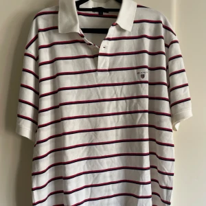 Gant pikétröja vitrandig stl XL - Vit pikétröja från GANT i storlek XL med horisontella ränder i marinblått och rött. Klassisk krage och korta ärmar, GANT-logga broderad på bröstet. Tröjan är gjord i mjuk bomull och har en normal passform.