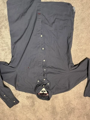 Mörkblå skjorta Tommy Hilfiger M - Snygg mörkblå skjorta från Tommy Hilfiger i slim fit med stretch. Klassisk design med knappar hela vägen och liten logga broderad på bröstet. Skjortan har långa ärmar och är gjord i mjuk bomull som är skön mot huden.