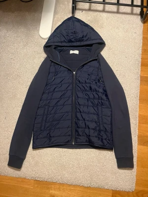 Mörkblå quiltad cardigan Pier One - Quiltad mörkblå cardigan från Pier One med huva och dragkedja framtill. Jackan har ribbade muddar vid ärmslut och snygga paneler i olika material. Perfekt för dig som gillar stilren och sportig look. Billigare vid snabb affär!