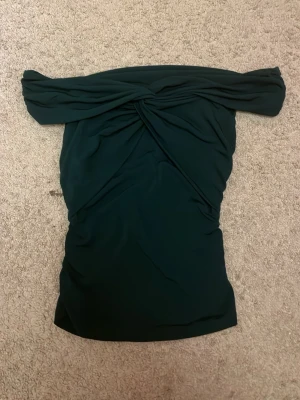 Mörkgrön offshoulder topp från Bikbok - Säljer en mörkgrön offshoulder topp från Bikbok med snygg twistad detalj framtill. Toppen är tight och har en axelbandslös design som ger en cool och trendig look. Materialet känns mjukt och stretchigt, perfekt för att styla med jeans eller kjol.
