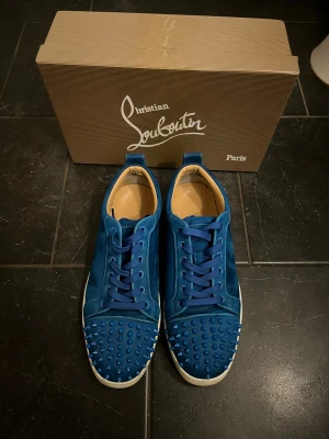 Blå Christian Louboutin med taggar - Säljer ett par blå sneakers från Christian Louboutin i mocka. Storlek 43, 3 taggar saknas på vänstra skon. Fortfarande i bra skick de ända är dem 3 saknade spikarna och att dem är smutsiga.