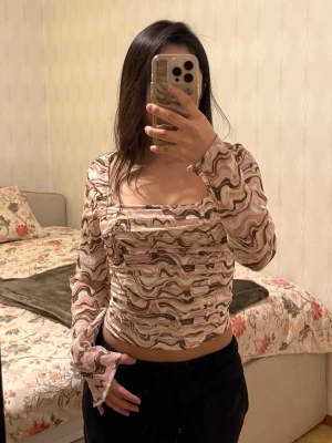 Mönstrad långärmad blus i pastell - Säljer en snygg blus med vågigt mönster i beige, rosa, brun och vit. Blusen har fyrkantig ringning, croppad passform och långa ärmar. Materialet är lätt och stretchigt, med smockad effekt på både fram- och baksida. Perfekt för dig som gillar unika prints och pastelltoner.