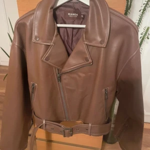 Bianca x nelly - Snygg brun skinnjacka med klassisk bikerstil från Bianca x Nelly är slut såld köpt för 699. Jackan har bred krage, asymmetrisk dragkedja, två sidofickor med dragkedja och ett matchande bälte i midjan. Perfekt för dig som vill ha en cool och tidlös look.Väldigt stori storleken