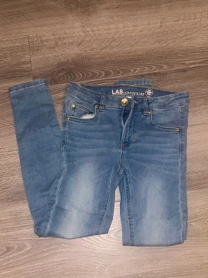 Jeans från LAB Industries, strl 134 - Stretchiga jeans från LAB Industries i storlek 134. Klassisk femficksmodell med guldfärgade nitar och knapp, tillverkade av 77% bomull, 21% polyester och 2% elastan.