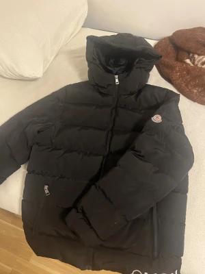 Svart Moncler pufferjacka med huva - Svart pufferjacka från Moncler med huva och dragkedja framtill. Jackan har Moncler-logga på ärmen och två sidofickor med dragkedja. Materialet är polyester och dun, perfekt för kalla dagar. Snygg och stilren design som håller dig varm hela vintern.