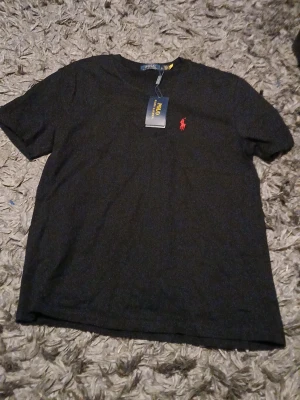Svart t-shirt från Polo Ralph Lauren - Svart t-shirt från Polo Ralph Lauren med klassisk röd logobrodering på bröstet. T-shirten har rund hals och korta ärmar, tillverkad i mjuk bomull för skön känsla. Perfekt för dig som gillar stilrena och enkla plagg med en touch av lyx.
