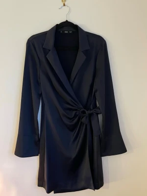 Svart omlottklänning  - Snygg svart kort omlottklänning från MNG med krage och långa ärmar. Klänningen har en  knytning i midjan och är gjord i sidenliknande material.