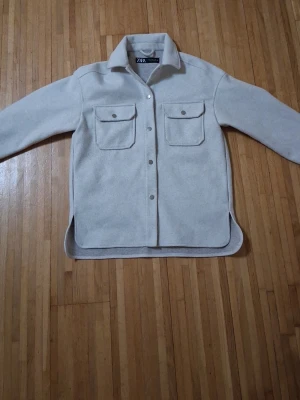 Ljusgrå overshirt från Zara - Stilren ljusgrå overshirt från Zara med två stora bröstfickor och knappar framtill. Overshirten har klassisk krage, långa ärmar och rundade slitsar i sidorna. Tillverkad i ett mjukt, filtliknande material som ger en clean och modern look. Använt bara ett par gånger.