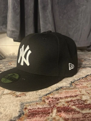 Svart New Era Yankees keps - Svart keps från New Era med New York Yankees-logga i vitt framtill och broderad Frihetsgudinna i blått och vitt på sidan. Klassisk flat peak och broderad New Era-logga på sidan. Perfekt för dig som gillar streetwear och sportig stil.