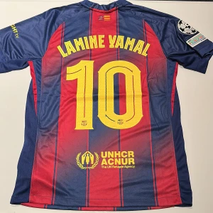 FC Barcelona Lamine Yamal #10 tröja Nike - Säljer en FC Barcelona fotbollströja från Nike med Lamine Yamal och nummer 10 på ryggen. Tröjan är blå och röd med gula detaljer, randigt mönster, Champions League-patch på ärmen och sponsorer som Spotify och UNHCR. Tillverkad i lätt och ventilerande polyester. Replika, endast handtvätt gäller! Defekter enligt sista bilden!