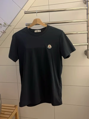 Mörkblå Moncler t-shirt med logga - Snygg mörkblå t-shirt från Moncler med klassisk rund halsringning och korta ärmar. På bröstet sitter Monclers ikoniska logga som en patch. Perfekt för en clean och stilren look.