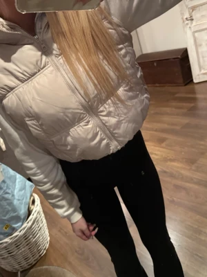 Beige cropped dunväst med huva - Snygg beige dunväst i cropped modell med dragkedja. Västen har två stora fickor framtill och en stilren, minimalistisk look. Perfekt att slänga över en hoodie för en trendig streetstyle.