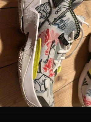 Jordan Why Not Zer0.4 Graffiti sneakers - Unika Jordan Why Not Zer0.4 sneakers med graffiti-inspirerat mönster i vitt, svart, rött och gult. Skorna har mesh- och textilmaterial, vita snören och coola detaljer som synliga sömmar och färgade inslag. Perfekta för dig som vill sticka ut med din stil. Open for discussing price 