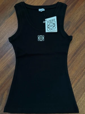  Loewe topp - Snygg svart ribbad linnetopp från Loewe med broderad vit logga framtill. Toppen är ärmlös och har en klassisk rund halsringning. Perfekt att styla med jeans eller kjol för en trendig look.