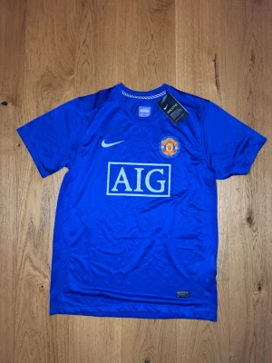 Blå Manchester United tröja Nike - Snygg blå Manchester United matchtröja från Nike med AIG-logga på bröstet och klubbmärke. Tillverkad i lätt och ventilerande material, perfekt för fotboll eller träning. MUFC-tryck i nacken och klassisk Nike Swoosh på framsidan.