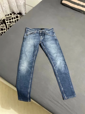Blå Tiger of Sweden jeans - Snygga blå slim jeans från Tiger of Sweden med klassisk femficksmodell och slitningar framtill. Jeansen har smal passform och detaljer i silverfärgad metall. Perfekta för en trendig och avslappnad look.