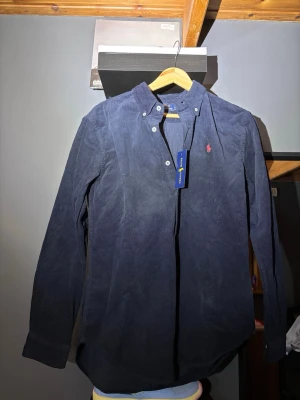 Mörkblå manchesterskjorta från Polo Ralph Lauren - Snygg mörkblå manchesterskjorta från Polo Ralph Lauren med klassisk krage och knappdetaljer. Skjortan har lång ärm och den ikoniska röda logotypen broderad på bröstet. Perfekt för dig som gillar stilrena och tidlösa plagg.