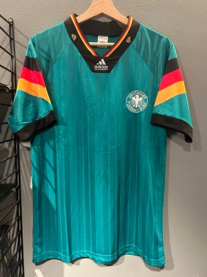 Tysklands retro fotbollströja Adidas - Grönblå retro fotbollströja från Adidas med Tysklands landslagsemblem på bröstet. Tröjan har svarta, röda och gula detaljer på ärmarna och svart krage med ränder. Tillverkad i glansigt syntetmaterial, perfekt för fotboll eller streetwear. Helt ny med tagg en replika 