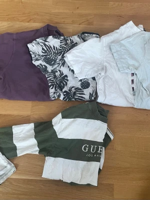 Fyra trendiga t-shirts i olika färger + 1 långärmad - Paket med fyra t-shirts: en lila, en vit med svart bladmönster, en helvit och en grön/vit randig från GUESS Los Angeles. Perfekta för en avslappnad och cool stil. Alla har korta ärmar och klassisk passform.