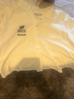 Vit skjorta från NOCCO Barebells - Vit skjorta med krage och knappstängning framtill från NOCCO Barebells. Skjortan har en bröstficka och tryckt logga på vänster sida. Perfekt för en avslappnad och stilren look. Storlek passar nog storlek S