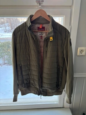 Olivgrön pufferjacka från Parajumpers - Snygg olivgrön pufferjacka från Parajumpers med quiltad front, ribbade muddar och dragkedja. Jackan har två fickor med dragkedja och en gul detalj vid kragen samt ett patch på ärmen. Perfekt för kalla dagar och riktigt stilren.