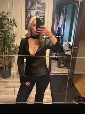 Svart omlottopp med djup v-ringning - Snygg svart i satin topp  med djup v-ringning och lång ärm. Toppen har choker-detalj o halsen. Helt ny med prislapp kvar. Passar S/M