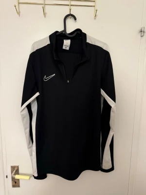 Svart Nike träningsset med vita detaljer - Säljer ett svart träningsset från Nike med vita detaljer på ärmar och axlar. Setet består av en långärmad tröja med halv dragkedja och matchande byxor. Perfekt för träning eller uppvärmning. Nike-logga på både tröja och byxor. 