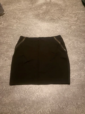 Svart kortkjol med dragkedjor - Snygg svart kortkjol med dragkedjor framtill vid fickorna. Kjolen har en enkel och stilren design som passar till många olika looks. Perfekt att matcha med en cool t-shirt eller stickad tröja.