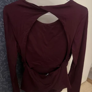 Vinröd topp från Gina tricot - Trendig vinröd långärmad topp med öppen rygg och snygg twistad detalj baktill. Tillverkad i mjukt material som sitter skönt mot kroppen. Perfekt för dig som vill ha en unik och stilren look.